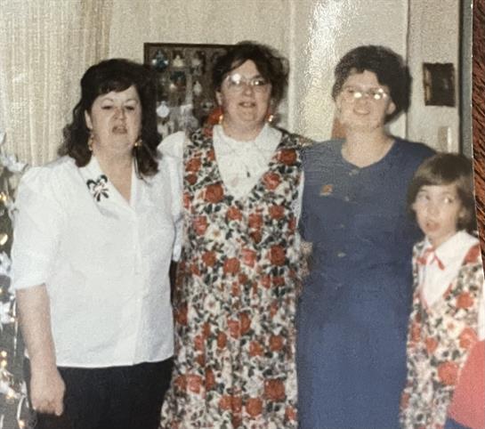 Four women and a girl smile together at a family gathering, dressed in colorful floral attire.