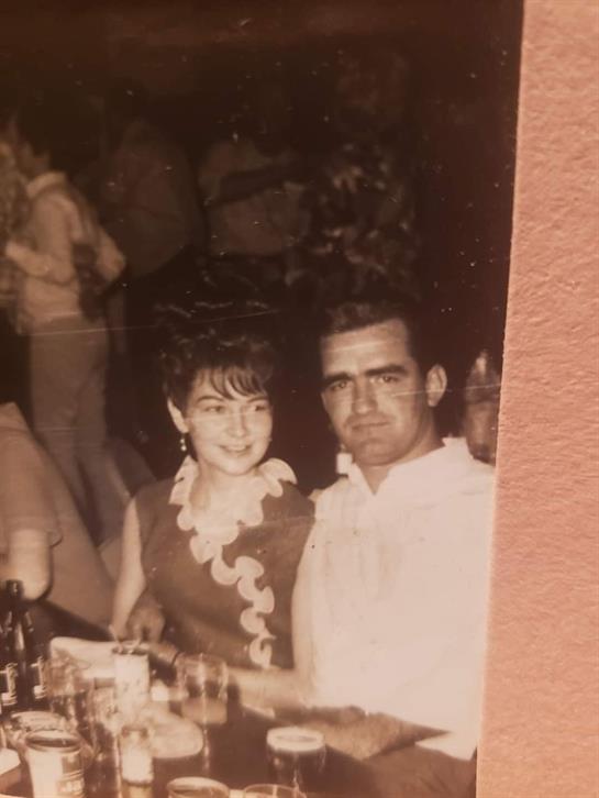 A couple shares a joyful moment at a crowded social event in the 1960s, surrounded by friends.