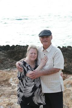 An elderly couple stands close together on a rocky beach, smiling happily as the sun sets.