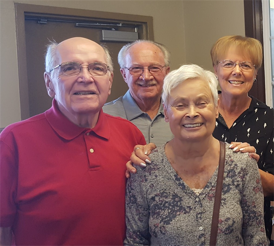Four elderly friends smile warmly, gathered closely in a cheerful indoor space.
