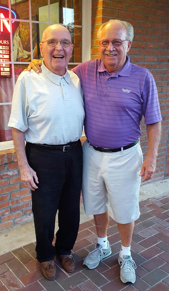 Two elderly men smile broadly while posing for a picture outside a brick restaurant.