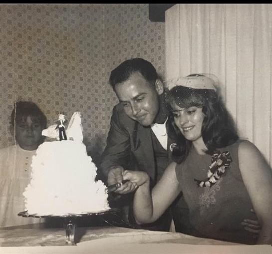 A couple smiles while cutting their wedding cake, with a guest watching in a vintage space.