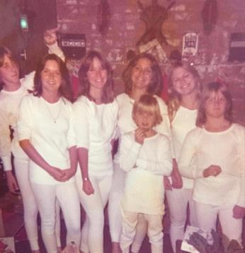 A group of six young women and two children dressed in white poses cheerfully in a cozy setting.
