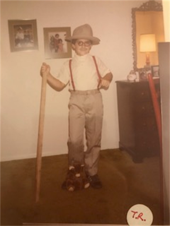 Child wearing oversized glasses and a hat stands confidently with a cane indoors.
