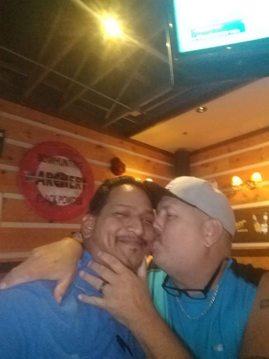 Two men embrace and smile in a bustling bar, capturing their joyful friendship in a playful kiss.