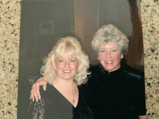 Two women are joyfully posing with arms around each other in a warm gathering setting.