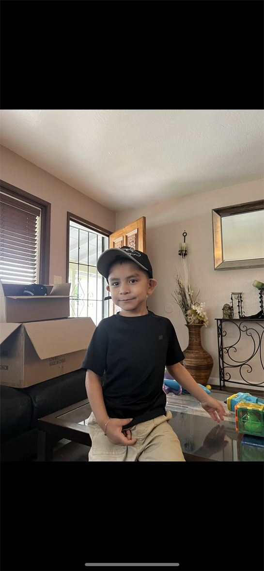 A boy in a black shirt and cap sits on a table in a living room filled with toys and boxes.