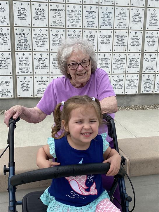 A cheerful elder and a smiling child share a moment near commemorative plaques in bright daylight.