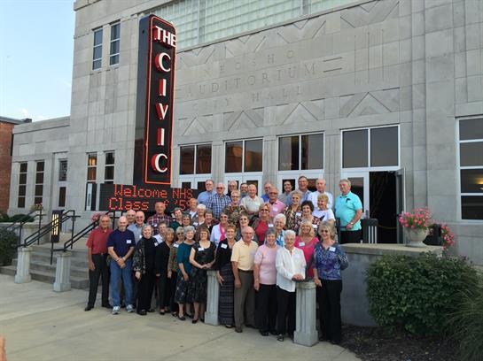 Numerous seniors pose together outside a civic center, celebrating a reunion under twilight.