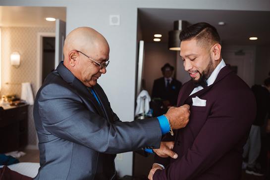 A father assists his son in adjusting his lapel in a stylish hotel room, preparing for an event.