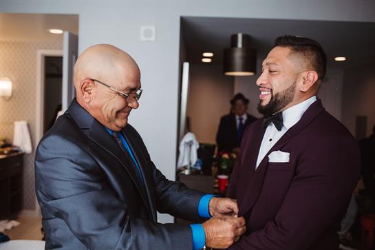 Groom smiles while father assists with tuxedo adjustments in a hotel room before his wedding.