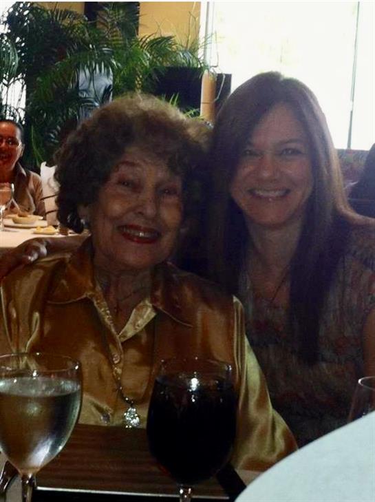 Two women smile while seated at a dining table with drinks and food during a lively gathering.