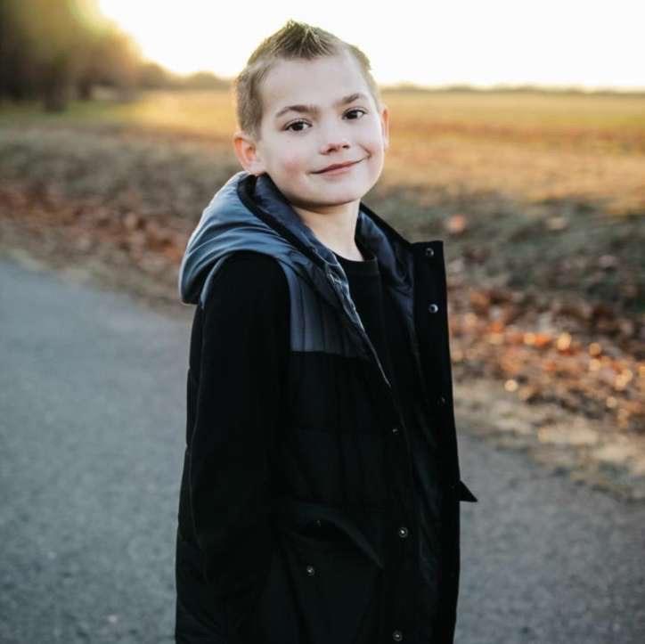 A boy poses confidently on a rural road during sunset, surrounded by fields and autumn leaves.