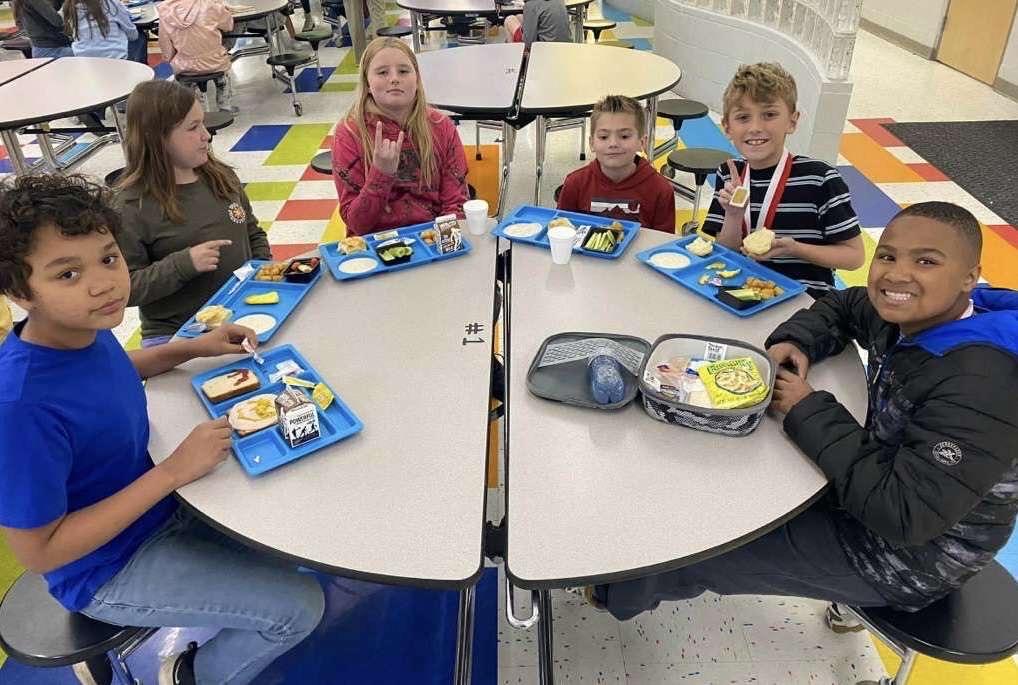 A group of children gather around tables to eat lunch, chatting and sharing food during break.