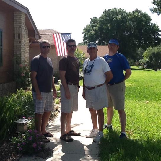 Four men stand on a sidewalk in a green yard, enjoying a sunny day together near a house.