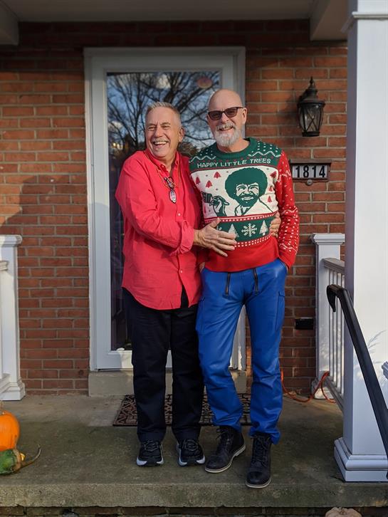 Two individuals smile while standing on a porch, enjoying a cheerful moment together.