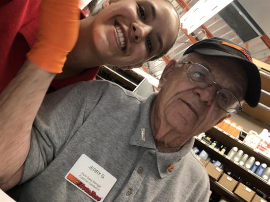 A young worker smiles broadly next to an elderly customer in a retail setting.