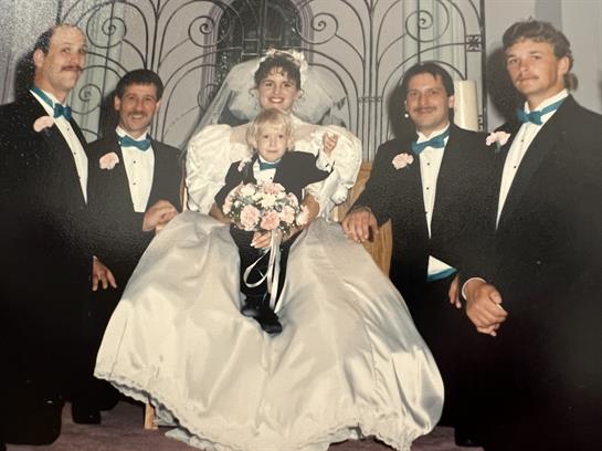 A bride in a stunning gown sits joyfully among her groomsmen and a little girl with flowers.