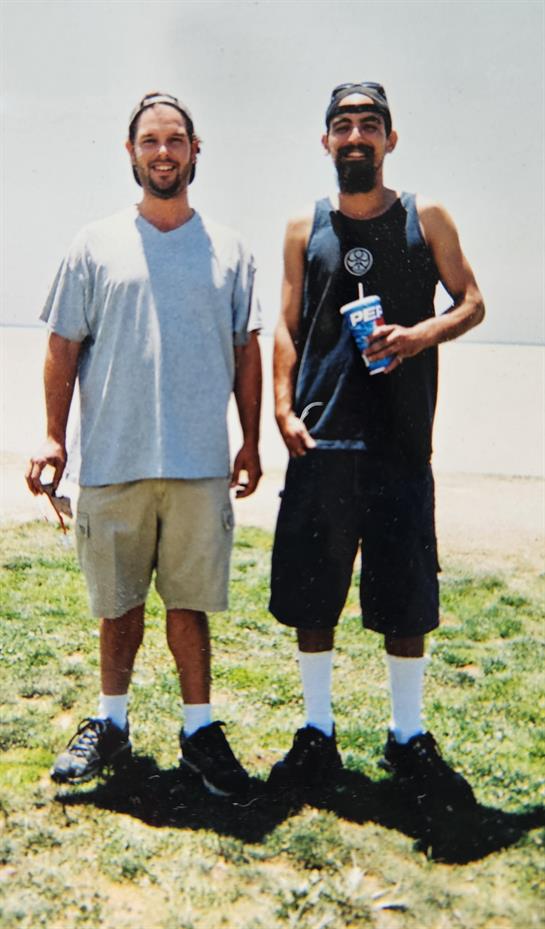 Two friends smile with drinks on grass by water on a sunny day.