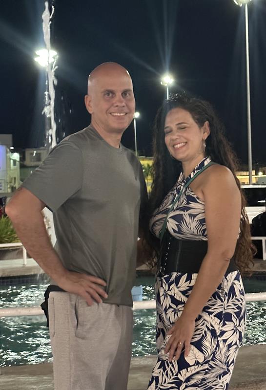 A couple smiles and poses together by a decorative fountain at night, dressed for a summer outing.