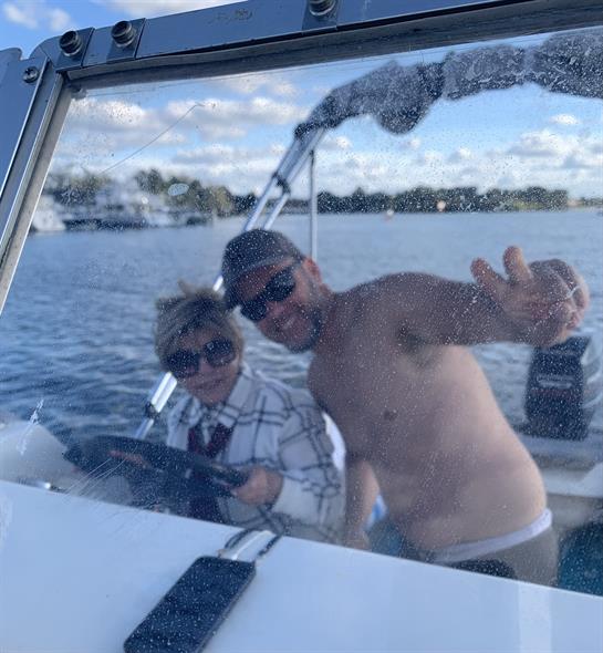 A father and son smile joyfully while steering a boat on a sunny day by the water.