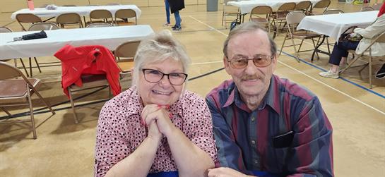 A smiling couple sits at a table, enjoying a community gathering in a spacious gymnasium.