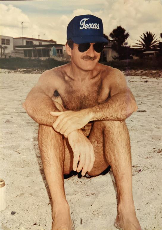 A shirtless man sits on the beach, wearing sunglasses and a cap, enjoying the warm weather.
