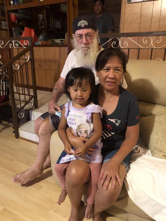 A child sits on the lap of an adult while another adult smiles nearby in a warm home setting.