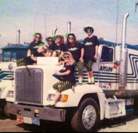 A group of women in matching outfits smiles on a classic truck under clear skies.