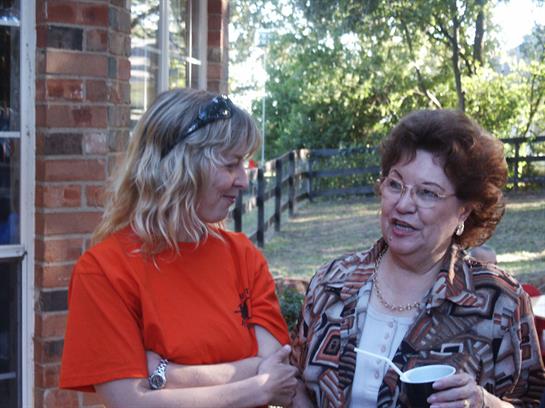 Two women smile and chat while enjoying a casual outdoor gathering in a green area.