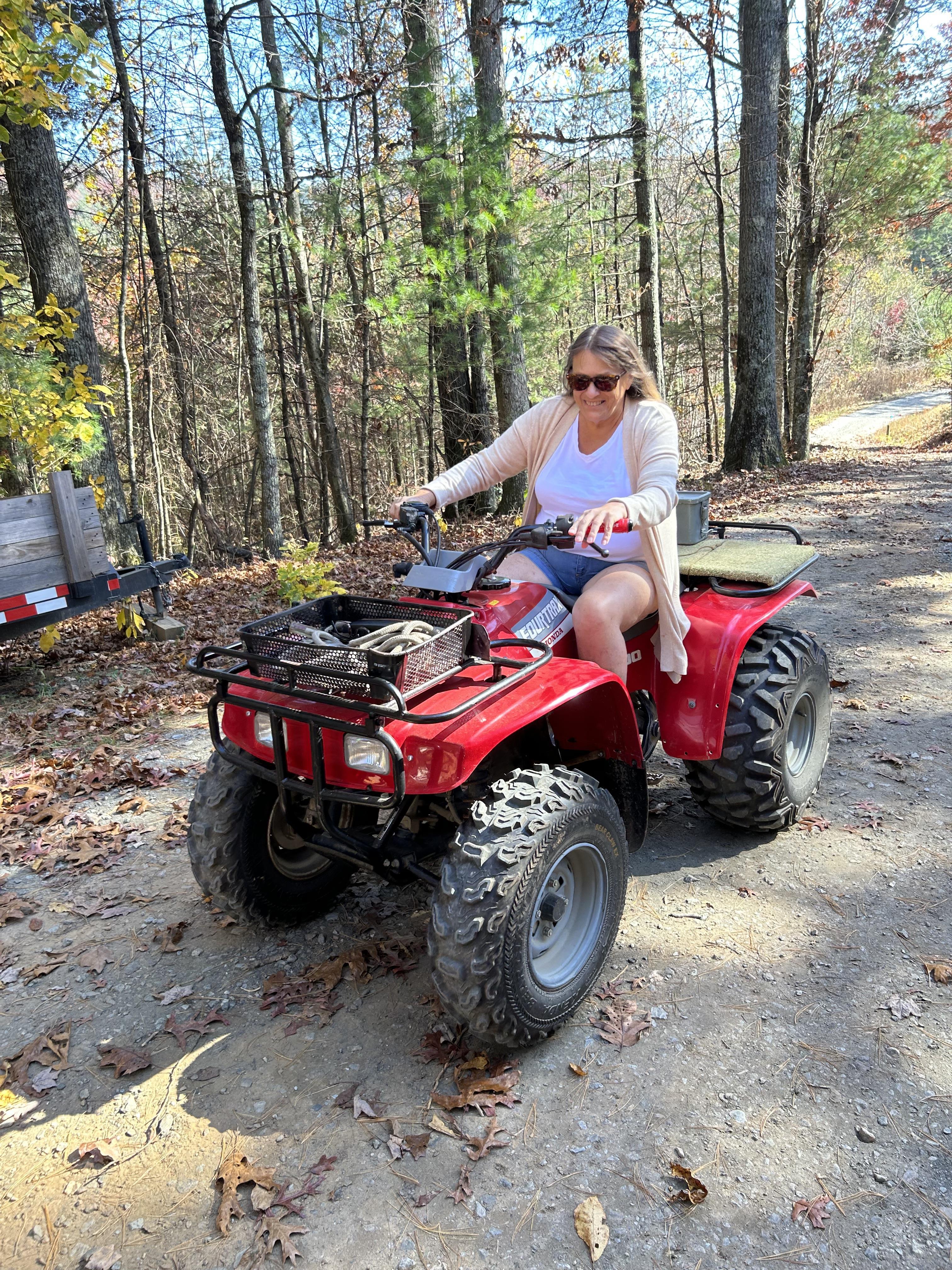 A woman drives an ATV along a dirt path surrounded by trees during a sunny fall day.