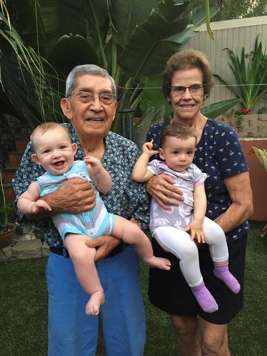 An elderly couple smiles broadly while holding their two grandchildren in a vibrant garden setting.