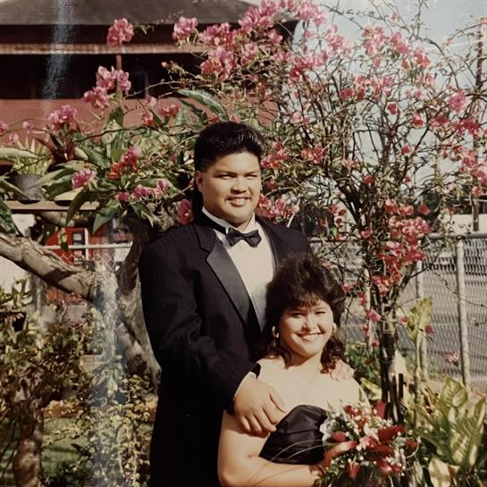 A couple stands together smiling in formal clothing surrounded by vibrant flowers in a garden.