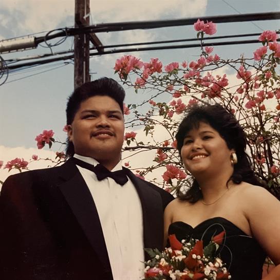 A happy couple poses together in formal wear, surrounded by vibrant pink flowers under a blue sky.