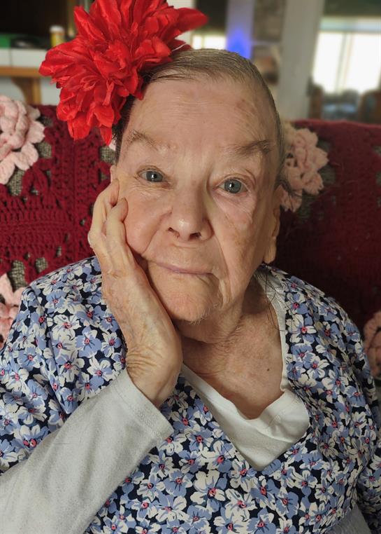 An elderly woman with a flower in her hair rests her chin on her hand, looking thoughtfully.