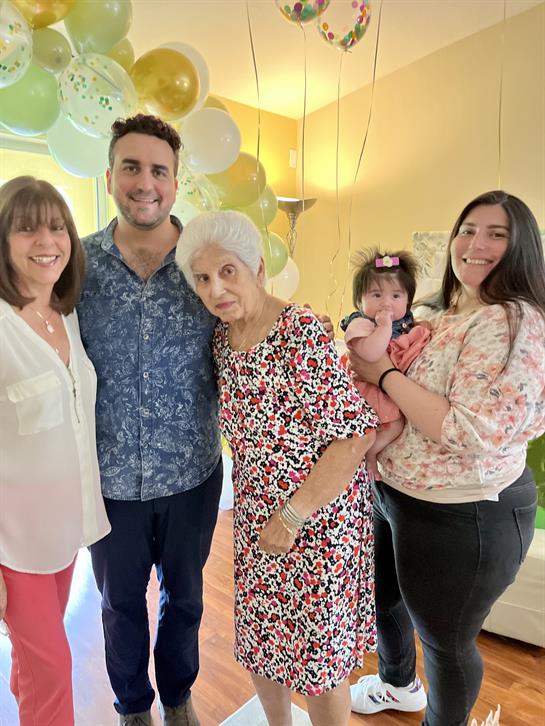 Five family members, including an elderly woman and a baby, celebrate together in a sunny room.