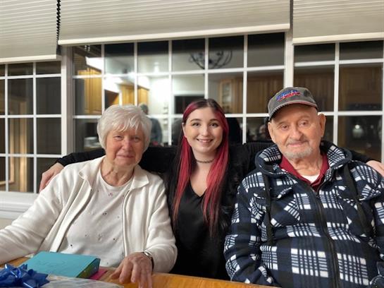 A granddaughter smiles between her grandparents in a cozy indoor setting during a family visit.