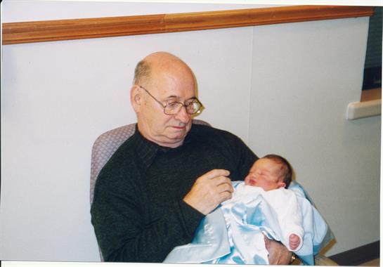 An elderly man cradles a newborn in a cozy chair, sharing a tender moment indoors.