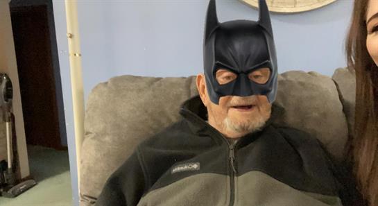 An elderly man wearing a batman mask sits relaxed on a couch while a young woman poses beside him.