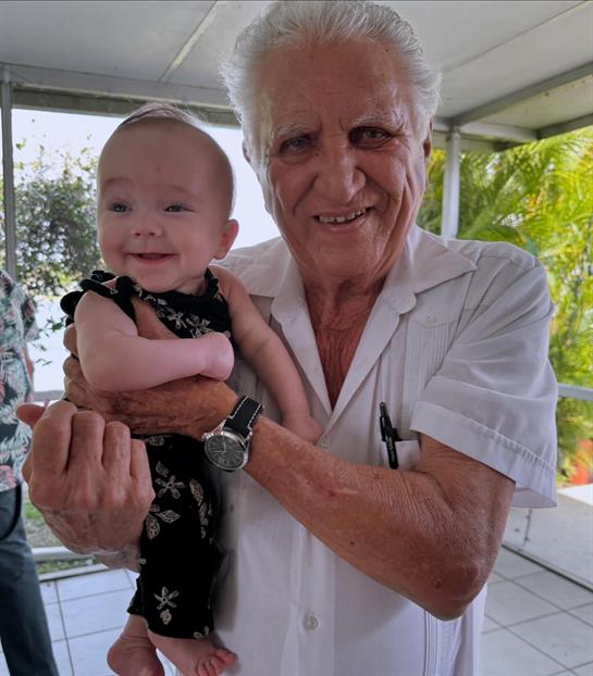 An elderly man cradles a happy baby girl in a warm, bright room filled with greenery.