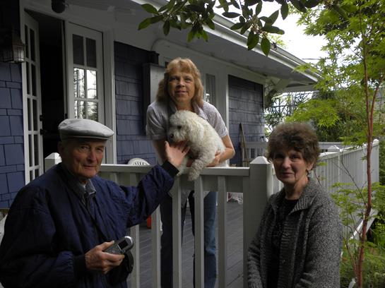 Three adults pose on a porch with a small white dog, smiling during an overcast afternoon.
