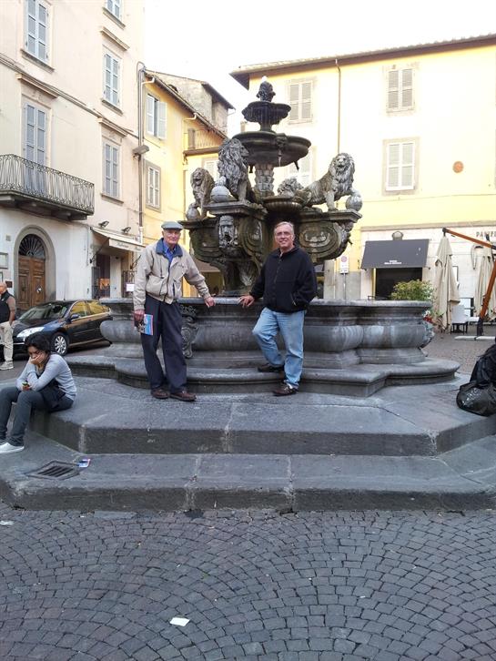 Two men stand at an intricately designed fountain while a person sits nearby in a lively square.