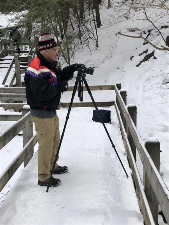 A photographer sets up his camera on a tripod amidst a snowy path in a forest.