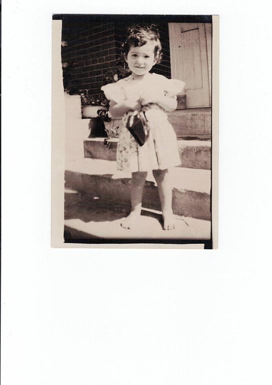 A cheerful young girl stands barefoot on a doorstep, clutching a small purse, exuding innocence.