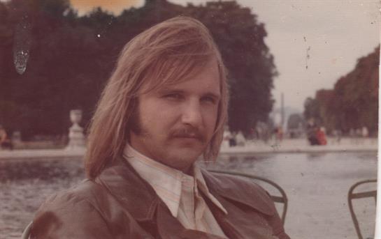 A man with long hair and a mustache enjoys a quiet moment by a pond in a park.
