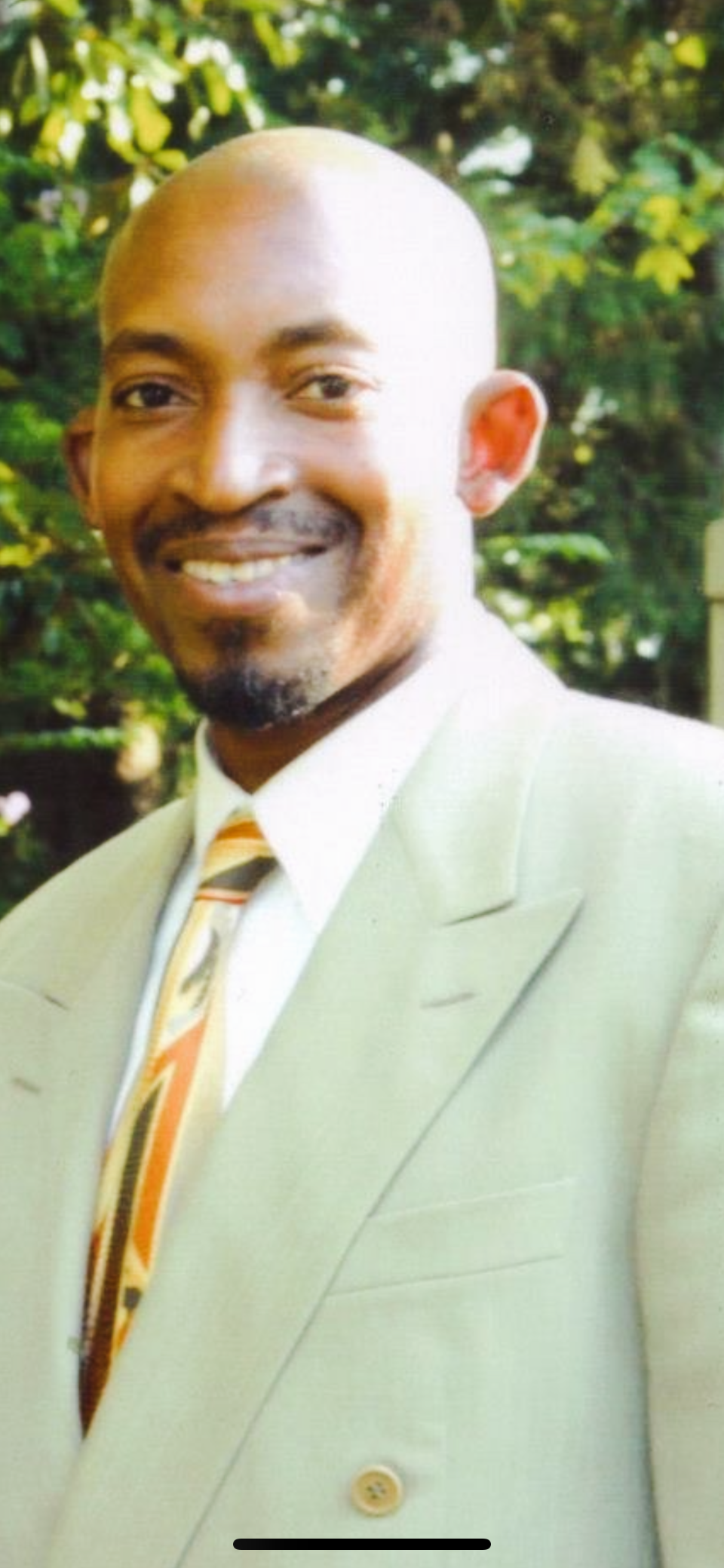 A man with a bald head and a wide smile stands outdoors, dressed in a light suit and colorful tie.