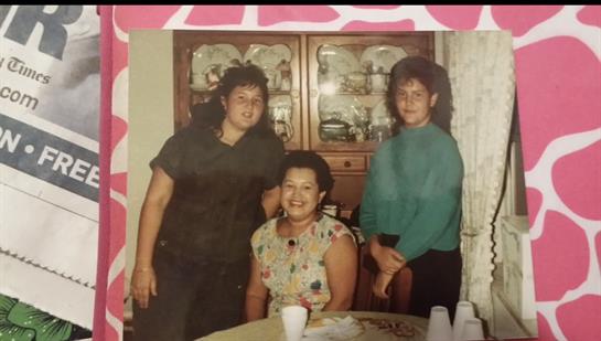 Three women are smiling and posing together in a warm living room filled with vintage decor.