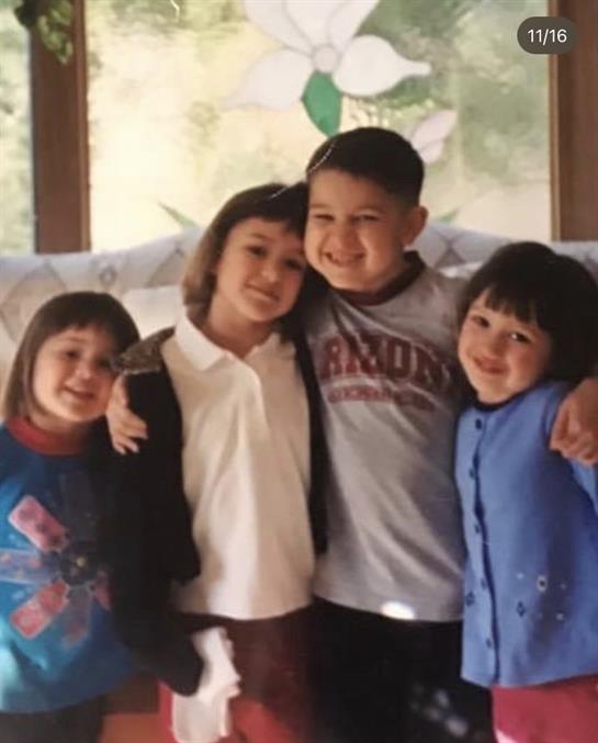 Four children stand together, smiling joyfully in a warm indoor environment.