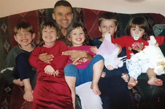 A joyful adult sits with five cheerful children on a couch, enjoying playful moments.