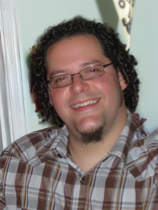 Man with curly hair and glasses smiling warmly while socializing with friends in a cozy setting.